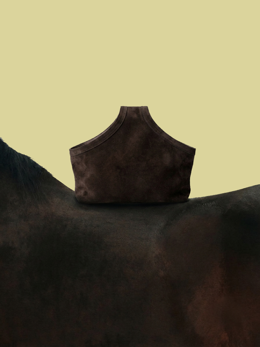 A dark brown suede or nubuck shoulder bag with a structured, asymmetrical slouchy silhouette, resting on the dark fur of a horse against a solid pale yellow background.